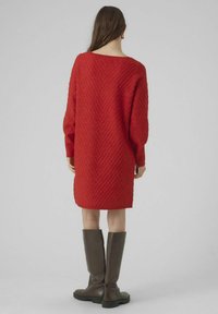 Robe en maille rouge avec un motif en chevrons, manches longues et encolure bateau, associée à des bottes marron montant jusqu'au genou.