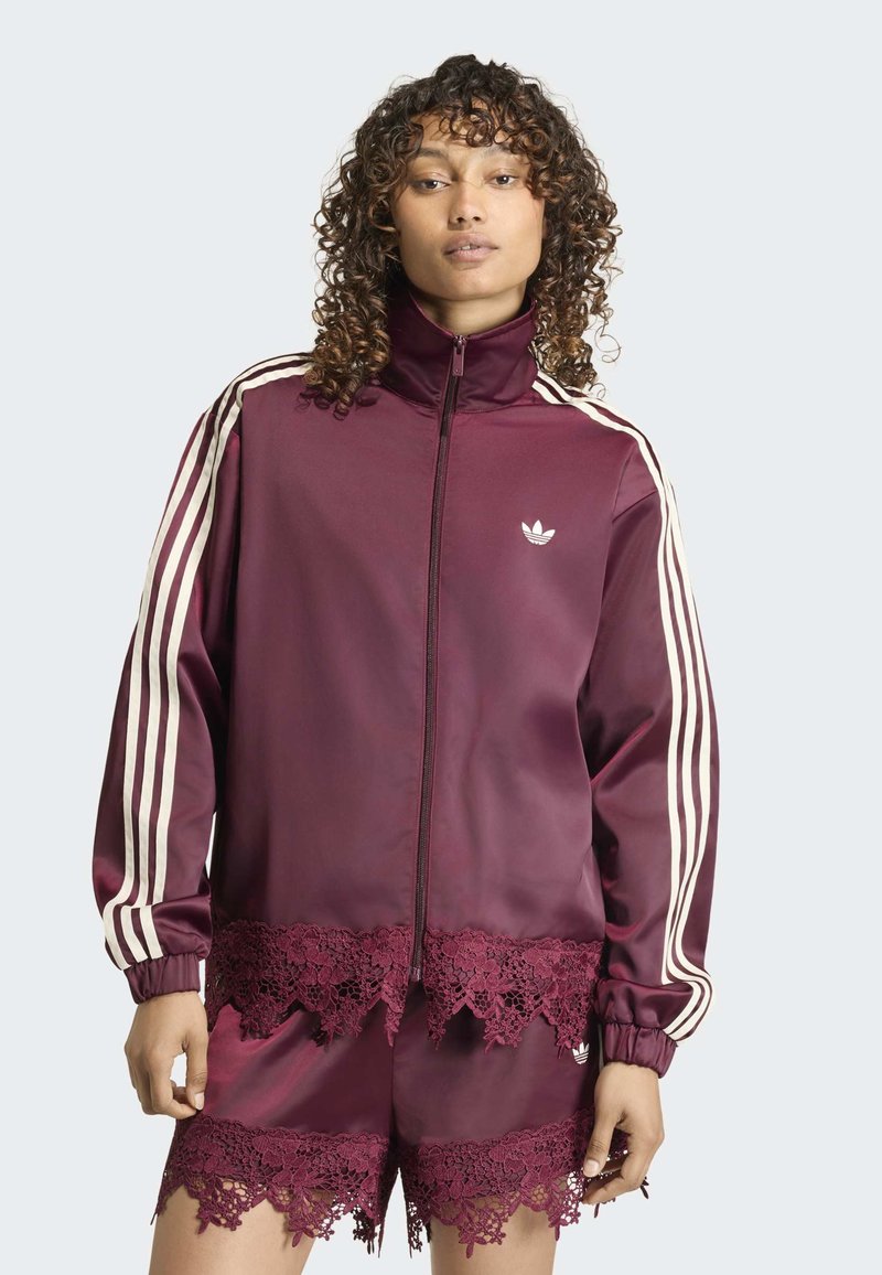 Femme aux cheveux bouclés portant une veste Adidas bordeaux et un short assorti avec une bordure en dentelle et des bandes blanches sur les manches.