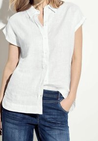 Camicia bianca di lino a maniche corte con frontale a bottoni e vestibilità morbida, abbinata a jeans denim scuri.