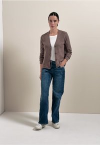 Brun stickad kavaj över en vit t-shirt, ihop med blå vida jeans och vita sneakers. Kavajen har en knapp-up framtill.