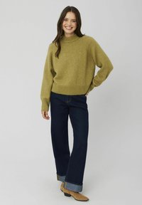 Pull en tricot vert olive avec un col montant et un ourlet côtelé, associé à un jean large bleu foncé et des bottines marron.