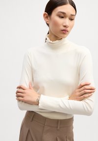 Hvid langærmet turtleneck top lavet af blødt, strækbart stof, med en flæset høj hals og tætsiddende ærmer, båret med beige bukser.