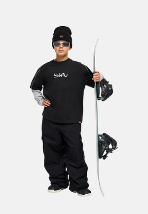 Junge Person in schwarzer Snowboardausrüstung und Mütze hält ein Snowboard aufrecht, trägt eine Sonnenbrille.