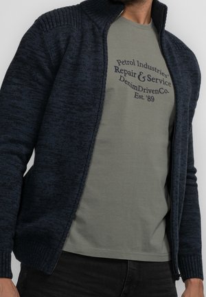Homme portant un sweat zippé bleu foncé par-dessus un t-shirt gris avec un texte noir indiquant "Petrol Industries Repair & Service DenimDrivenCo. Est. '89".