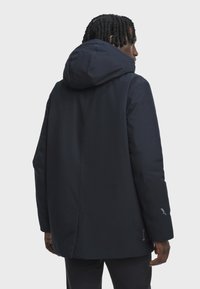 Man wearing a black hooded GORE-TEX jacket facing away, showing back and right side against a plain light gray background.