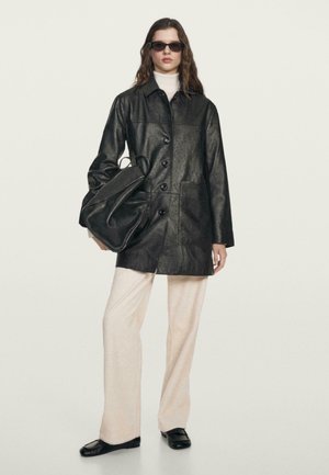 Giacca nera in finta pelle con bottoni, tasche frontali, sovrapposta a un dolcevita chiaro. Abbinata a pantaloni chiari e a una borsa nera, design elegante.