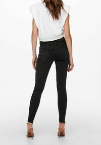 Jeans negros ajustados de cintura alta, con bolsillos traseros y una textura suave. Combinados con una blusa blanca y tacones de color claro.