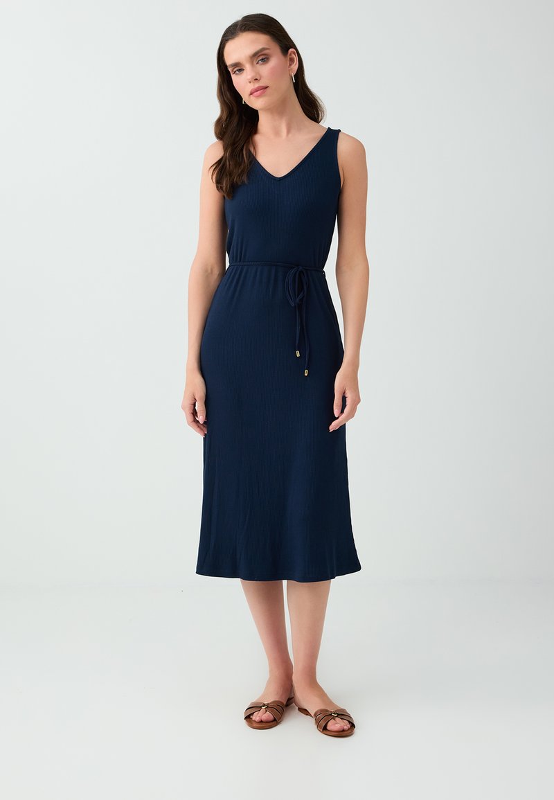 Femme debout portant une robe midi sans manches bleu marine avec une ceinture nouée à la taille et des sandales plates marron sur un fond clair uni.