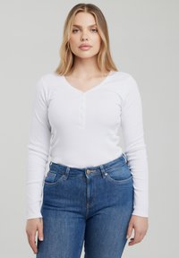 Femme blonde portant un haut henley blanc à manches longues et un jean taille haute bleu, debout devant un fond gris clair uni.