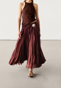 Jupe maxi plissée bordeaux à l'apparence texturée, associée à un haut marron sans manches ajusté et des sandales noires à lanières.