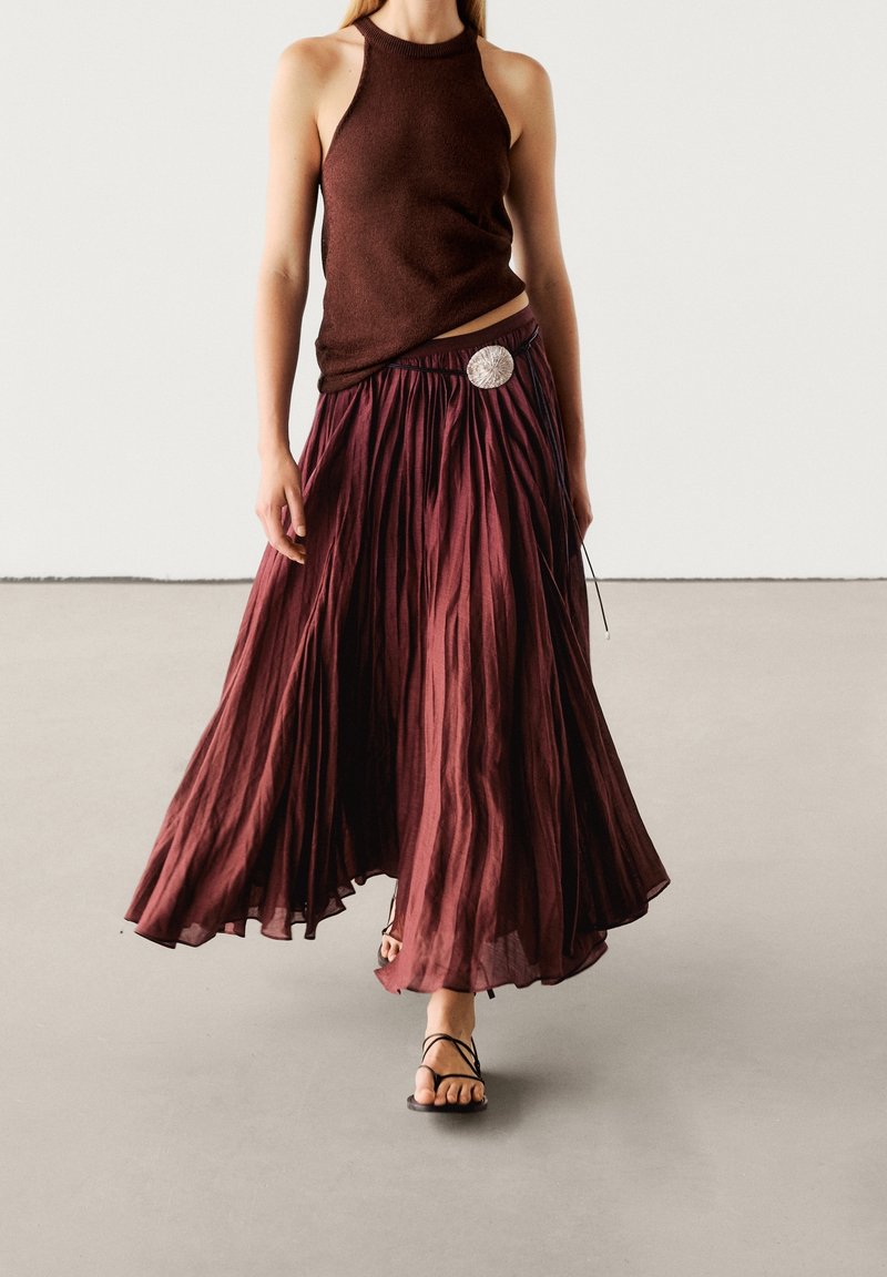 Jupe maxi plissée bordeaux à l'apparence texturée, associée à un haut marron sans manches ajusté et des sandales noires à lanières.