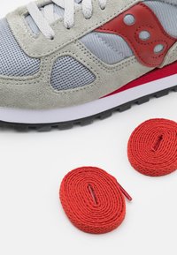 Sneaker grigia con texture in rete e suede, caratterizzata da accenti rossi e una suola intermedia bianca. Include due lacci rossi arrotolati.