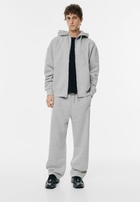 Siva dukserica s prednjim zatvaračem i vezicom, u kombinaciji s usklađenim trenirkama. Ispod je crna majica. Crne tenisice upotpunjuju ovaj outfit.