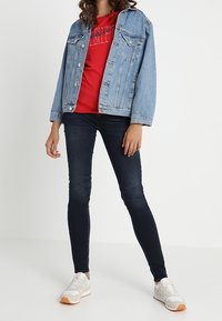 Mujer con chaqueta de mezclilla azul claro, camiseta gráfica roja, jeans ajustados oscuros y zapatillas blancas, de pie sobre un fondo blanco.