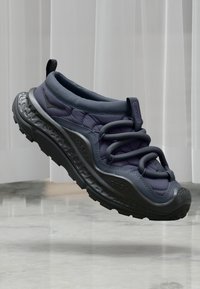 Ciemnoszare i czarne sneakersy z grubą podeszwą i pętlami sznurowadeł, unoszące się nad odblaskową marmurową podłogą, przed pionowymi żaluzjami.