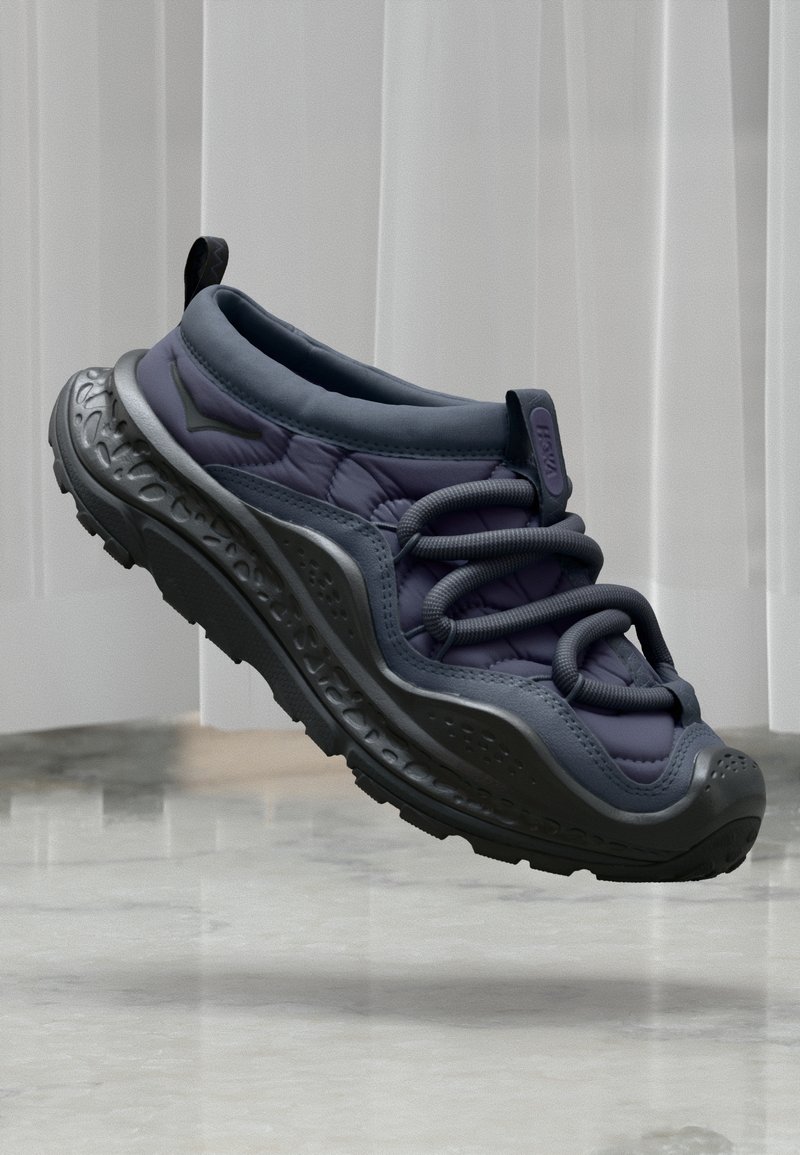 Ciemnoszare i czarne sneakersy z grubą podeszwą i pętlami sznurowadeł, unoszące się nad odblaskową marmurową podłogą, przed pionowymi żaluzjami.