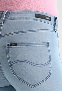 Jeans de mezclilla azul claro con un corte ajustado. Las características incluyen una etiqueta de cuero con "Lee" en la cinturilla y bolsillos traseros curvados.