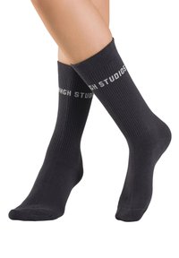 Schwarze Baumwollsocken mit Rippstrickstruktur und weißem "AHGH STUDIO"-Schriftzug am oberen Rand. Mittellange Länge bis zur Wade, glatte Oberfläche.