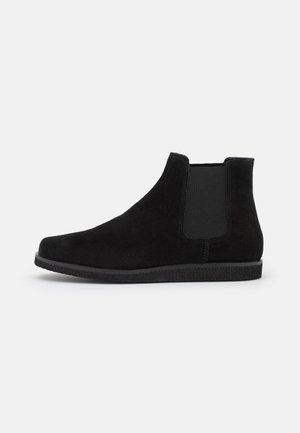 Bota Chelsea de ante negro con panel lateral elástico y suela plana de goma, mostrada desde el lado exterior sobre un fondo blanco.