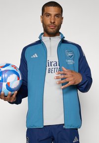 Blau und marinefarbene Zip-Jacke mit Adidas-Logo, Arsenal-Wappen und grauem Hemd darunter. Hält einen mehrfarbigen Fußball.