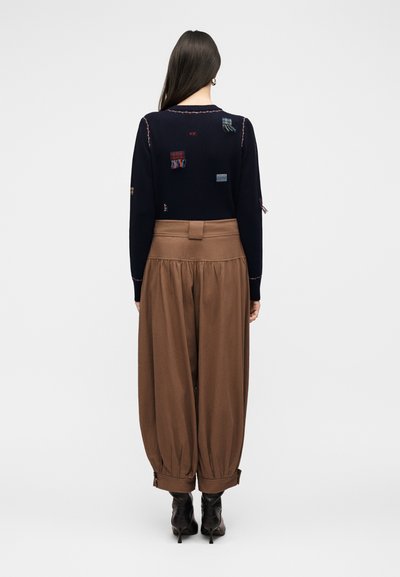 Γυναίκα με μακριά σκούρα μαλλιά που φοράει μπλε σκούνο πουλόβερ με καρό μπαλώματα και φαρδιά καφέ παντελόνια που είναι μπαλωμένα μέσα σε μαύρες μπότες με τακούνι, στέκεται με την πλάτη στον φακό.