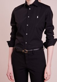 Person som bär en svart skjorta med knappar och uppvikta ärmar, svarta byxor och ett svart bälte med en guldkoppel, står mot en neutral bakgrund.