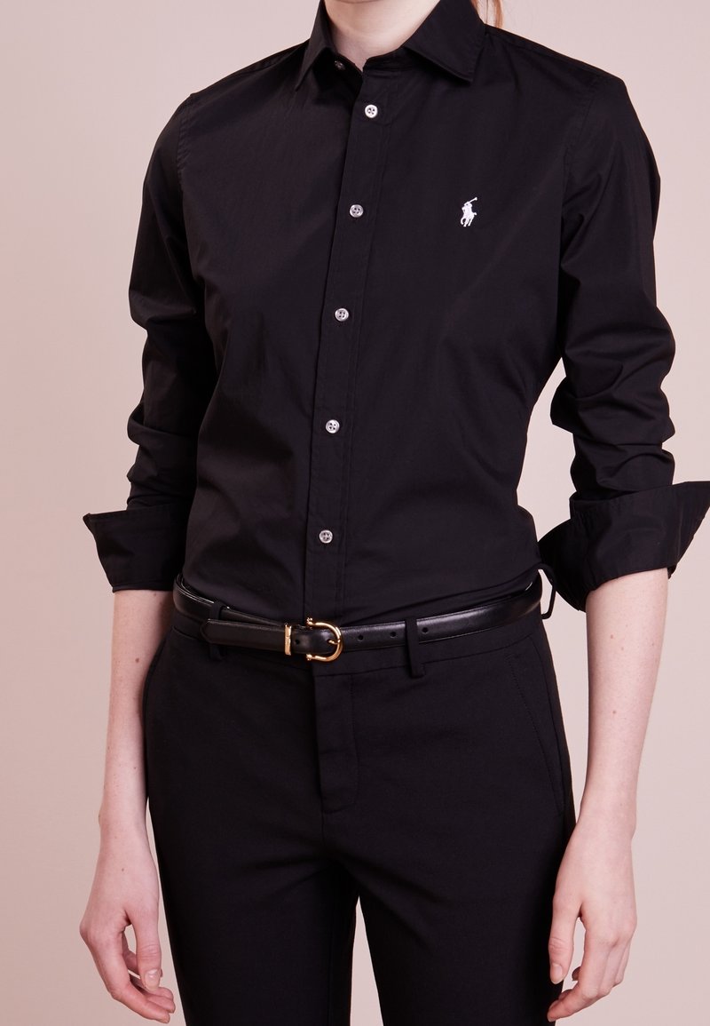 Person som bär en svart skjorta med knappar och uppvikta ärmar, svarta byxor och ett svart bälte med en guldkoppel, står mot en neutral bakgrund.