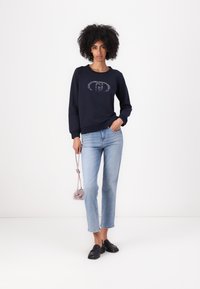Felpa blu navy con una grafica sulla parte anteriore, abbinata a jeans dritti in lavaggio chiaro e mocassini neri; tiene una borsa rosa.
