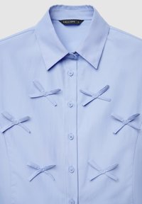 Camicia azzurro chiaro con sei fiocchi decorativi in tessuto disposti sul davanti e colletto standard.