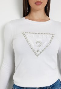 Maglietta bianca a maniche lunghe con un logo triangolare accentuato in argento e strass. Tessuto morbido con design aderente, abbinata a jeans in denim blu.
