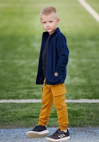 Chłopiec w granatowej kurtce na zamek, czarnym t-shircie i musztardowych spodniach typu cargo. Czarne sneakersy z białymi podeszwami. Proste uczesanie. Zielone tło.