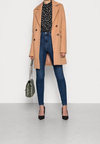Abrigo de mezcla de lana beige con botones de doble botonadura, blusa negra con lunares, jeans ajustados de tiro alto azules, tacones negros de punta, bolso verde.