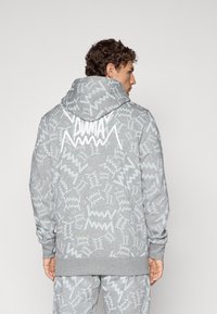 Mand iført grå Puma hættetrøje og matchende bukser med hvide abstrakte zigzag-mønstre, stående med ryggen til en ensfarvet baggrund.