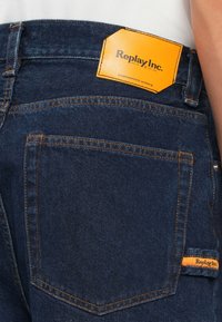 Mørkeblå denimjeans med oransje søm, som har et fremtredende oransje skinnmerke merket "Replay Inc." over en baklomme.