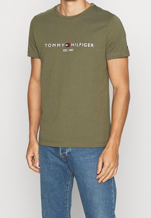 Mężczyzna w oliwkowej koszulce Tommy Hilfiger z logo i napisem "EST. 1985", w niebieskich dżinsach, stojący na jednolitym tle.