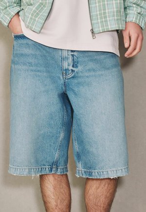 Mann trägt hellblaue Jeansshorts, ein weißes Hemd und eine grüne, kariert Reißverschlussjacke mit den Händen in den Taschen.