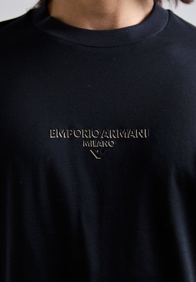Prim-plan al unei persoane îmbrăcate cu un tricou negru cu guler rotund, având logo-ul în relief "EMPORIO ARMANI MILANO" și emblema vulturului pe piept.