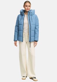 Kvinna som bär en ljusblå puffjacka med huva, krämfärgad tröja, vida krämfärgade byxor och vita sneakers med svarta ränder.