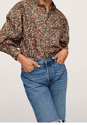 Person iført en langærmet blomstret bluse stoppet ned i højtaljede blå denimjeans med hænderne i forlommerne.