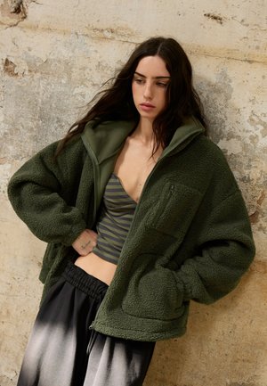 Jeune femme aux longs cheveux foncés portant une veste polaire verte par-dessus un crop top rayé, debout contre un mur beige texturé.
