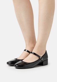 Black patent leather shoes with a rounded toe and a low block heel. Features a thin ankle strap with a silver buckle detail.