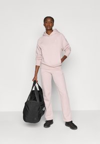 Haut à capuche rose clair et pantalon de survêtement assorti en tissu doux. Baskets de sport noires et sac de sport noir avec des sangles. Détail de logo simple.