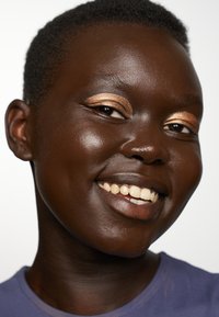 Gros plan sur une personne à la peau foncée, cheveux courts et fard à paupières doré, souriant. Elle porte une chemise violette, affichant une peau lisse et des dents blanches.