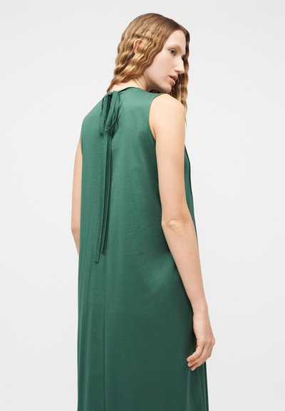 Γυναίκα με κυματιστά μαλλιά που φοράει πράσινο αμάνικο φόρεμα δεμένο στο πίσω μέρος, σε προφίλ σε απλό λευκό φόντο.