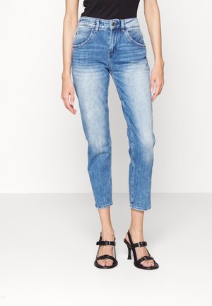 Femme portant un jean délavé taille haute bleu clair associé à des sandales à talons noires à bout ouvert, debout devant un fond blanc uni.