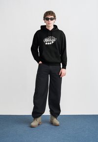 Μαύρο φούτερ με κουκούλα και λευκό λογότυπο, συνδυασμένο με φαρδιά μαύρη παντελόνια και γκρι μπότες. Το μοντέλο φοράει σκοτεινά γυαλιά ηλίου και στέκεται σε μπλε χαλί.