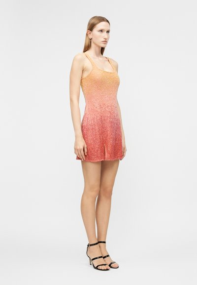 Γυναίκα στέκεται φορώντας αμάνικο, ροζ-πορτοκαλί πλεκτό φόρεμα με βαθμιαία απόχρωση και σκίσιμο μπροστά, μαζί με μαύρα σανδάλια με ψηλό τακούνι, σε απλό λευκό φόντο.