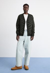 Cardigã de malha verde escuro com padrão zig-zag, t-shirt branca, jeans largos azul claro e sapatos de camurça castanho em fundo azul.