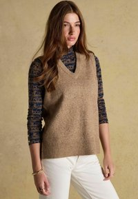 V-kaelusega, ilma varrukateta beež kampsun, millel on soonikkoes ääred, on kihitatud sinise pikavarukaga pluusi peale, millel on kuldne loomade muster. Valged püksid täiendavad komplekti.