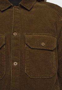 Camisa de pana marrón con un patrón de relieve texturizado, que presenta un cuello con puntas, dos bolsillos en el pecho y botones marrones.